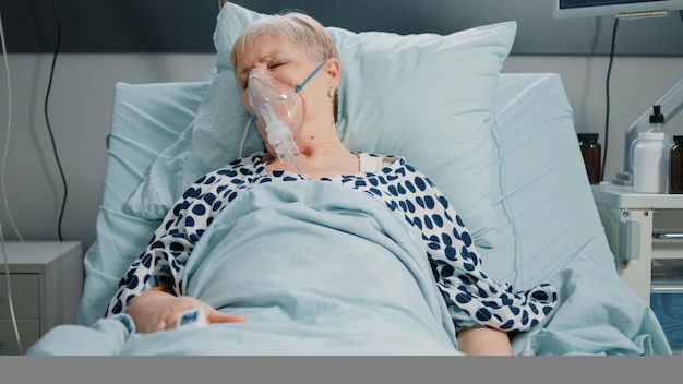 Retired woman with oxygen tube against respiratory problem in\
hospital ward bed. sick patient breathing heavily while resting and\
waiting for medical assistance with iv drip bag.
