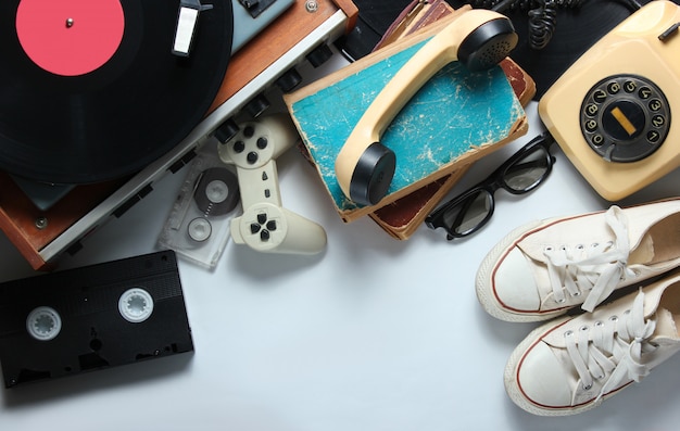 Retro 80s pop culture objects on white background. Copy space. Sneakers, rotary phone, vinyl player, old books, audio, video tapes, 3d glasses, gamepad.