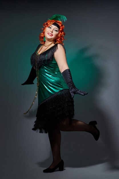 Retro cabaret plump woman with red hair in bright green dress on dark green background. Girl in Gatsby style dancing and posing. Russia, Sverdlovsk, February 14, 2018