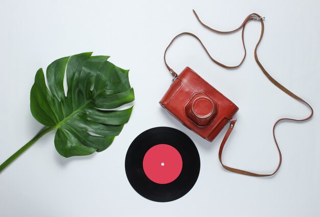 Retro camera in a leather case with strap, vinyl record and green monstera leaf on white background. Vintage flat lay style. Top view