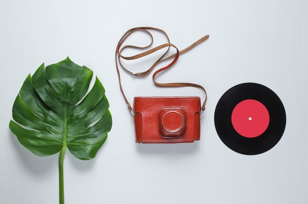 Retro camera in a leather case with strap, vinyl record and green monstera leaf on white background. Vintage flat lay style. Top view