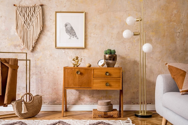 Retro composition of living room with wooden vintage commode, grey sofa, gold lamp, macrame, carpet, pillows, gold mock up poster frame, plants, decoration and personal accessories.