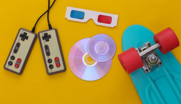 Retro entertainment, gadgets and devices. Gamepads, cruiser board, Cd's, 3D glasses on yellow background. 80s. Top view. Flat lay