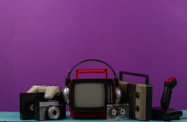 Retro portable mini tv set with headphones, radio receiver, joystick, gamepad, audio and video cassette, radio receiver on purple background. Attributes 80s, retro style