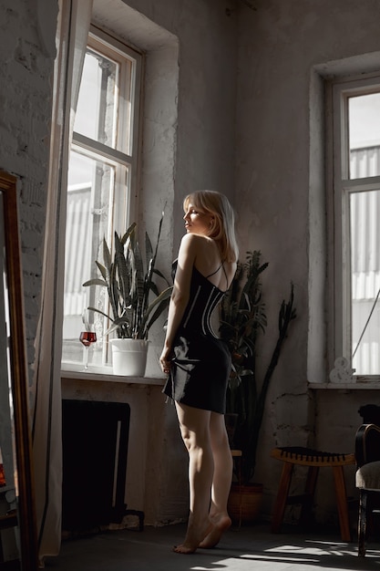 Retro portrait sexy blonde woman in black dress, vintage interior woman posing at window. Sensual romantic look, girl relaxes at home