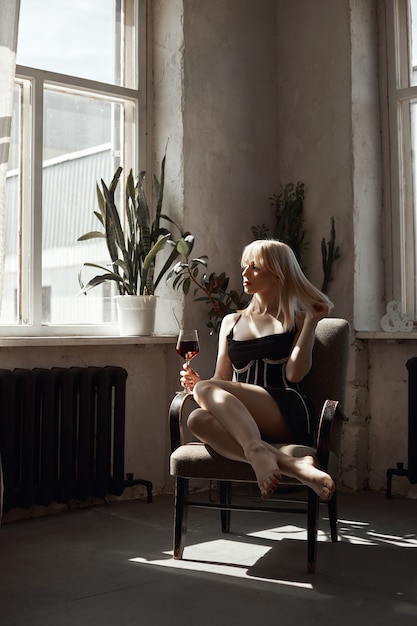 Retro portret sexy blonde vrouw met glas wijn in zwarte jurk, vintage interieur vrouw poseren bij raam. Sensuele romantische look, meisje ontspant thuis