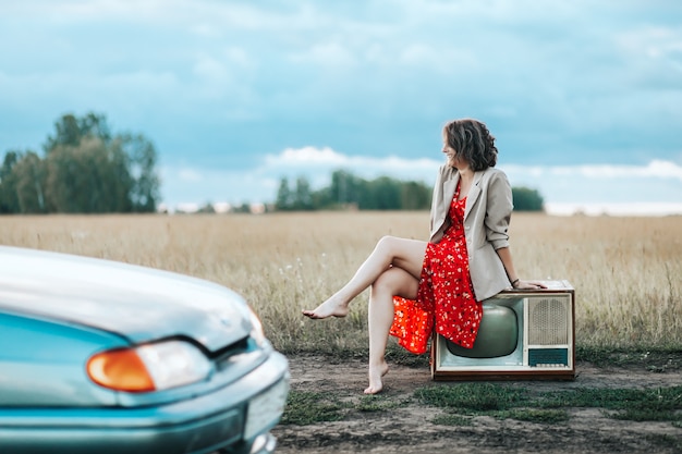 Retro portret van een jonge vrouw met een rode jurk en een beige jas zit op een oude tv