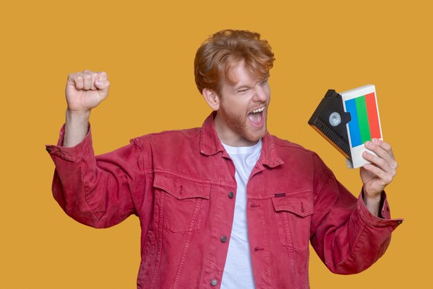 Photo retro. red-haired bearded young man in red jacket holding a video cassette