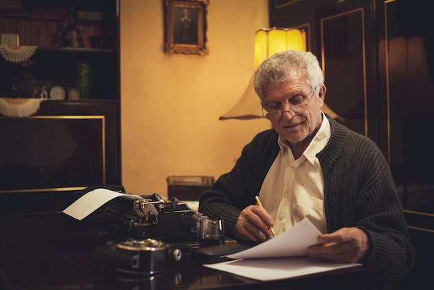 retro senior man writer with glasses is writing with pencil beside  obsolete typewriter.