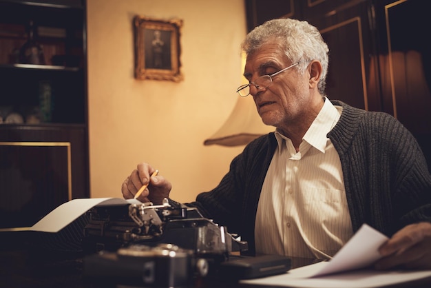 Foto scrittore uomo anziano retrò con occhiali e matita in mano seduto alla scrivania e leggendo del testo per scrivere su una macchina da scrivere obsoleta.