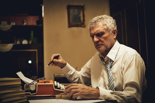 Retro senior man writer with pencil in his hand sitting at the desk and reading some text for writing on obsolete typewriter.