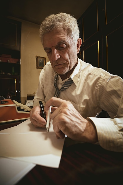 Retro senior man writer with pencil in one and cigarette in the other hand, sitting at the desk and writting some text on the paper.