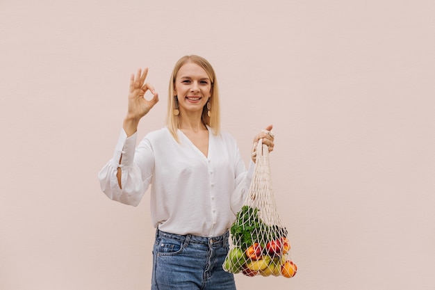 Reusable eco bag for shopping. Young woman holding string shopping bag with fruits and makes ok gesture