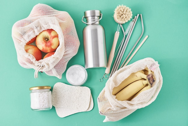 Reusable plastic free items on green surface. Glass jar, metal straws, aluminum bottle, wooden cleaning brush and fruits in shopping bag, top view