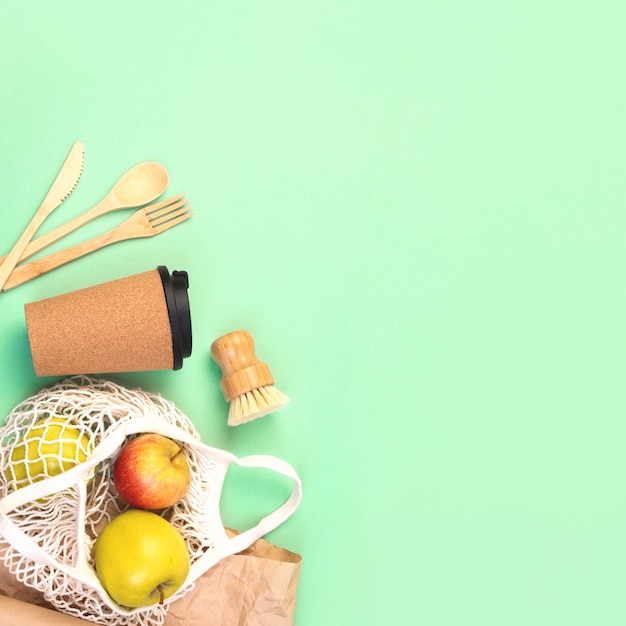 Photo reusable wooden cutlery, cork mug and grocery bag with apples. dishwashing brush and kraft wrapping paper, eco friendly fork, knife, spoon on green mint square backdrop. zero waste concept. copyspace.