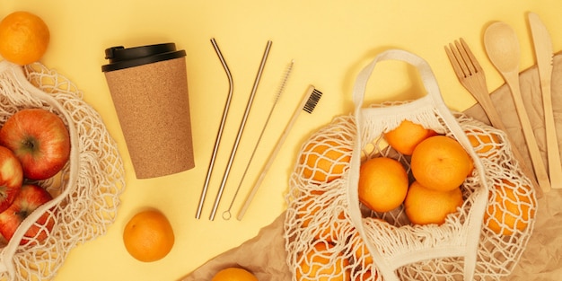 Photo reusable wooden cutlery, cork mug and grocery bag with fruits on yellow banner. zero waste concept.