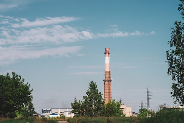 Reuze rookstapel op industriezone achter bomen. industriële structuur met grote pijp van bruine baksteen onder blauwe hemel. technologie en natuur. vervuiling van het milieu.