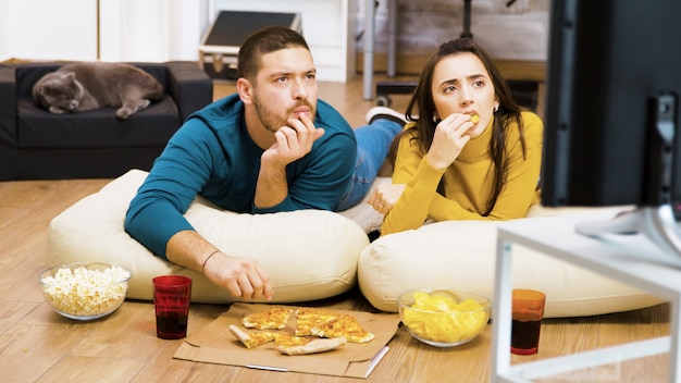Revealing shot of young couple watching tv sitting on pillow for the floor eating pizza while their cat is sleeping.