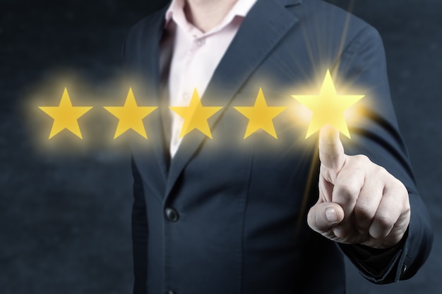 レビューと評価は会社のコンセプトを増やし、ビジネスマンの手は5つ星に触れます。評価と分類の概念。ビジネスマンは、5つの金の星をクリックして、会社の評価を上げます。