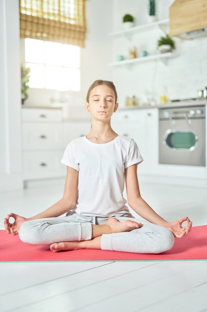 Revitalizing experience. Sporty relaxed hispanic teenage girl in sportswear looking calm while practicing yoga, sitting in Lotus pose on a mat in the kitchen. Home interior background