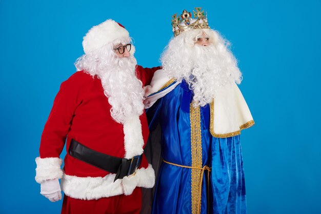 Rey mago and santa claus hugging as friends on a blue background