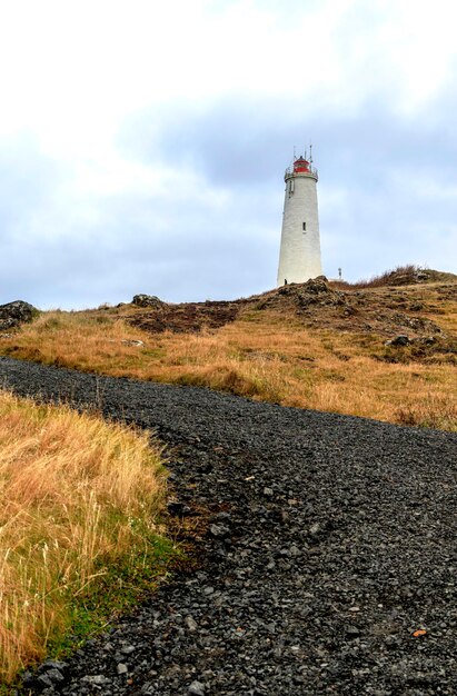 Маяк Рейкьянес Исландия Белый маяк, расположенный на вершине холма, окруженный желтыми травяными полями и грунтовой дорогой
