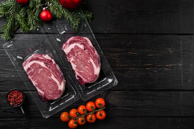 Rib-eye beef steak in plastic packing tray with christmas tree
decorations set, on black wooden table background, top view flat
lay, with copy space for text