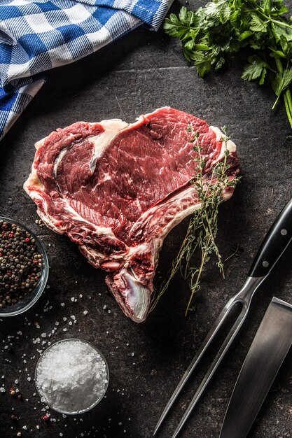 Rib eye steak with bone on butcher board with herbs salt pepper fork and knife.