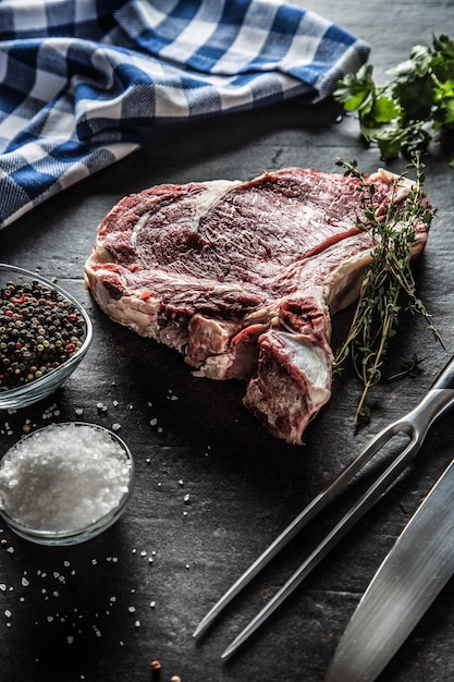 Rib eye steak with bone on butcher board with herbs salt pepper fork and knife.