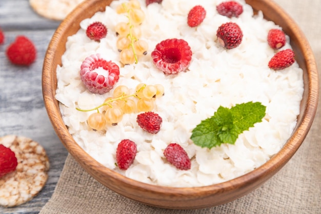 Rice flakes porridge with milk and strawberry in wooden bowl on gray wooden background and linen textile. Side view, selective focus, close up.