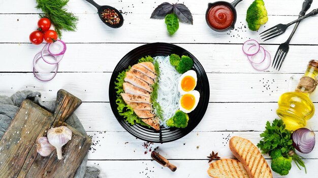 Rice noodles with chicken fillet egg and broccoli in a black plate on a white wooden background Top view Free space for your text