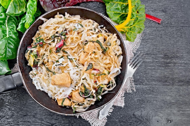 Rice noodles with leafy beet chicken breast meat cashew nuts and soy sauce in a frying pan on burlap on wooden plank background on top