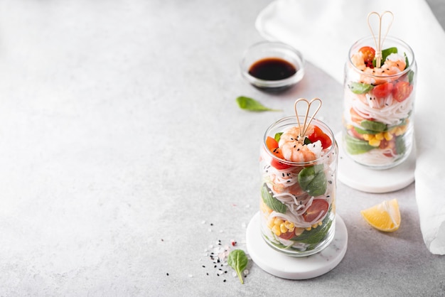 Rice noodles with shrimps and vegetables in a glass jar on a white table, top view