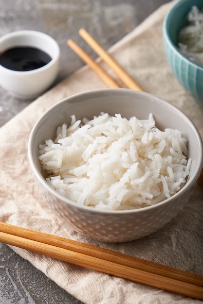 Рис в фарфоровой миске, с японскими палочками для еды, соевый соус, подается на сером каменном столе. Крупным планом.