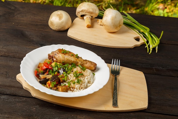 Rice with stewed vegetables and baked chicken leg on a handmade wooden board next to mushrooms and green onions