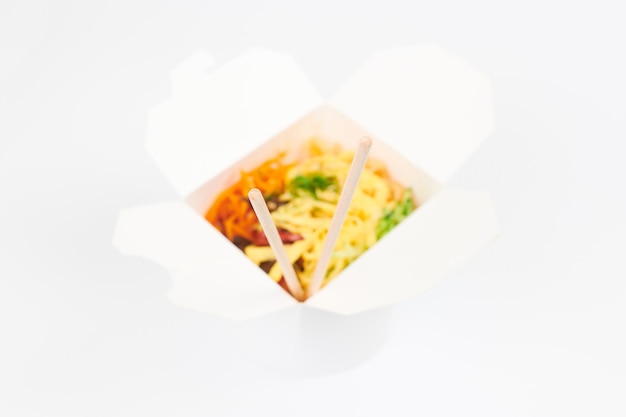 Rice wok with seafood and vegetables in white box and wooden chopsticks isolated on white background, top view, selective focus. Open takeout box with wok and bamboo sticks. Fast food delivery concept