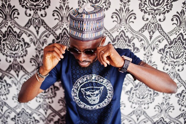 Rich african man in stylish traditional clothes and hat in glasses posed at studio against vintage background