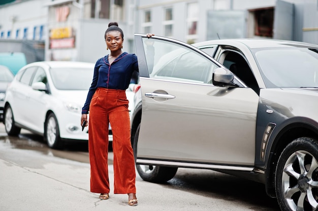 Donna africana d'affari ricca in pantaloni arancioni e camicia blu posata contro un'auto suv d'argento con la porta aperta