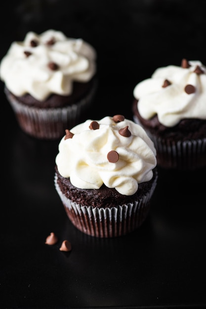 ホイップクリームのフロスティングとブラックの背景にチョコレートチップを添えたリッチなチョコレートカップケーキ