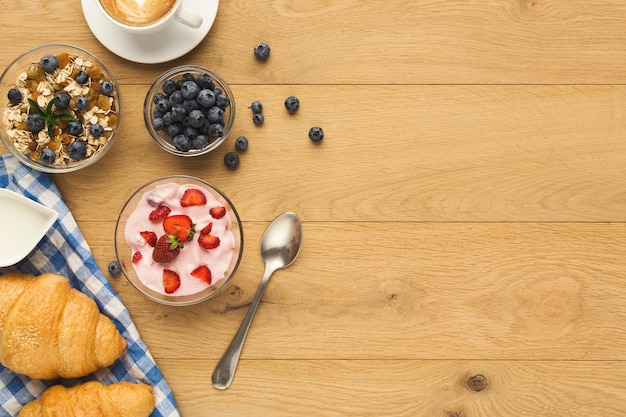 Rich continental breakfast background. French crusty croissants, muesli, yogurt, sweet berries and hot coffee for tasty morning meals. Delicious start of the day. Top view, copy space on natural wood