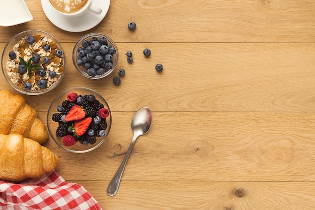 Rich continental breakfast background. French crusty croissants, muesli, yogurt, sweet berries and hot coffee for tasty morning meals. Delicious start of the day. Top view, copy space on natural wood