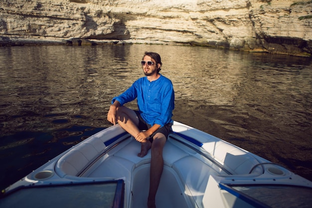 Rich free man in sunglasses and a blue shirt sits on the bow of white boat