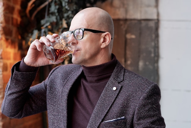Богатый лысый мужчина средних лет в очках стоит в ресторане-лофте и пьет алкоголь из стекла