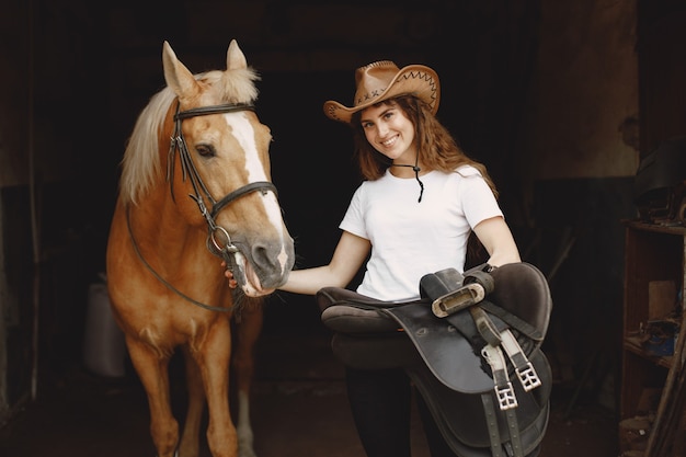 Foto donna del cavaliere che tiene una sella in una stalla. la donna ha i capelli lunghi e una maglietta bianca