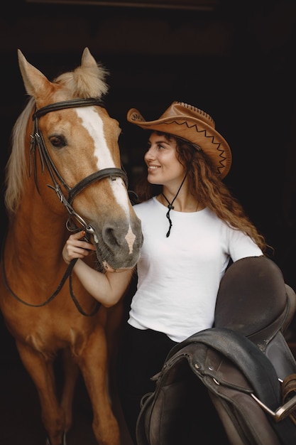 Foto donna del cavaliere che tiene una sella in una stalla. la donna ha i capelli lunghi e una maglietta bianca