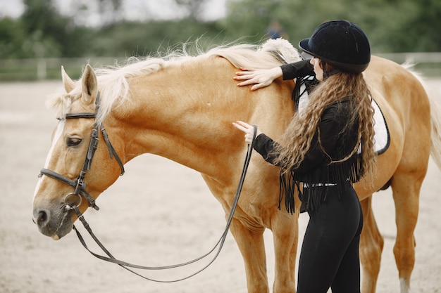 牧場で彼女の馬と話しているライダーの女性。女性は長い髪と黒い服を着ています。サドルに触れる女性の乗馬。