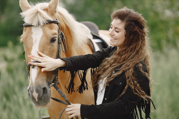 Rider woman talking to her horse on a ranch. Woman has long has and black clothes. Female equestrian touching her brown horse.