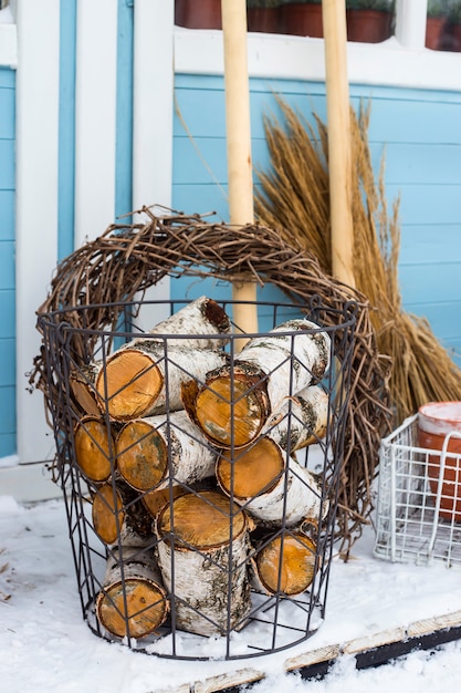 Rieten metaalmand met berkehout naast de tuinhulpmiddelen bij de muur van een blauw buitenhuis