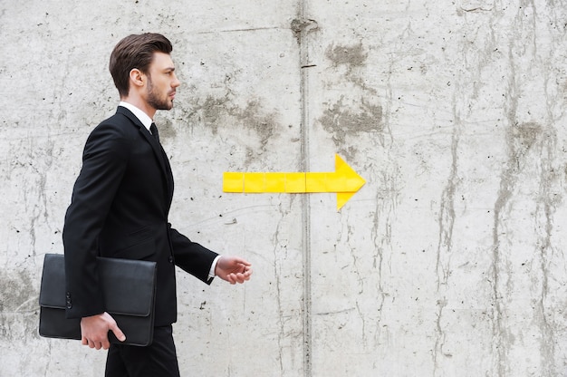 Right direction. Handsome young man in formalwear holding briefcase while walking in front of the concrete wall