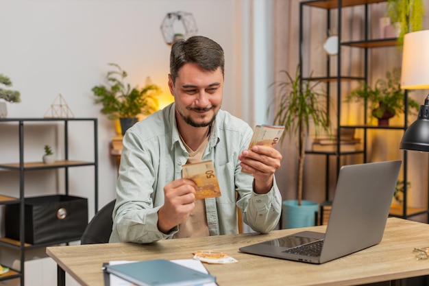 Rijke, gelukkige blanke man die geld telt, contant geld gebruikt met een laptop, huishoudelijke rekeningen berekent aan de tafel thuis.